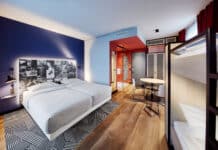 Ein Hotelzimmer, rechts steht ein Stockbett, im Zentrum ein Doppelbett mit hellgrauem Bezug. Die Wände sind in Blao, hellblau und rot gestrichen. Auf dem Boden liegt ein geometrisch gemusterter Teppich in schwarz-weiß.