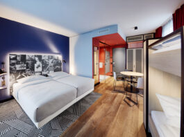 Ein Hotelzimmer, rechts steht ein Stockbett, im Zentrum ein Doppelbett mit hellgrauem Bezug. Die Wände sind in Blao, hellblau und rot gestrichen. Auf dem Boden liegt ein geometrisch gemusterter Teppich in schwarz-weiß.
