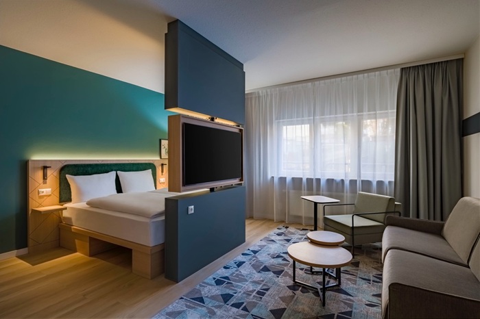 Ein Zimmer im neuen Sindelfinger Spark by Hilton mit einem Doppelbett hinter einer Trennwand, in der sich ein Fernseher befindet. Auf der anderen Seite ist ein Sofa.