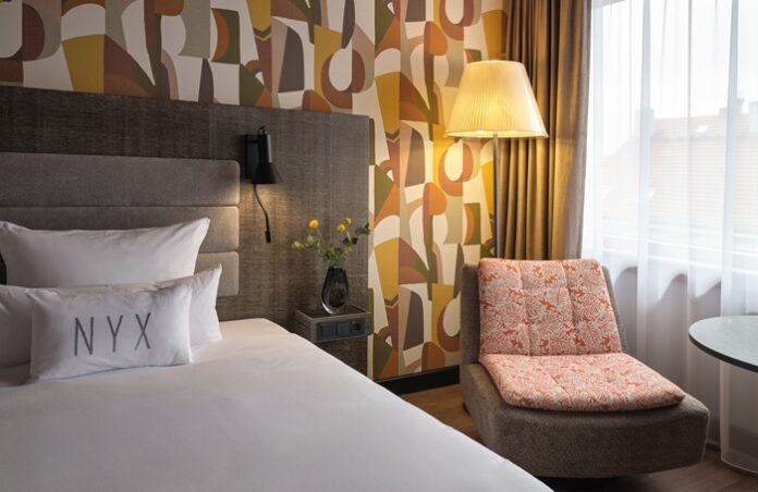 Hotelzimmer mit Bett und Stuhl vor grafisch gemusterter Tapete in Erdtönen
