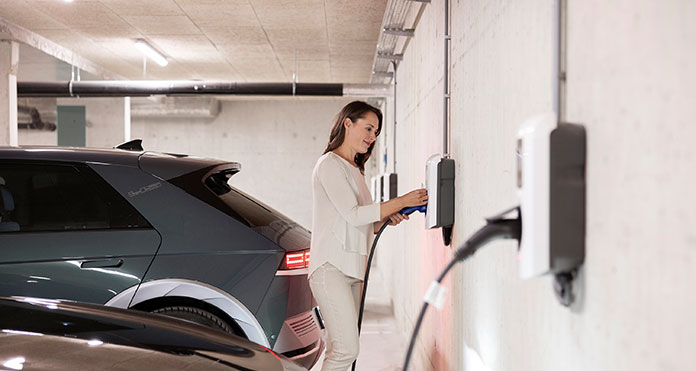 In einer Tiefgarage steckt eine Frau ein Ladekabel in eine an der Wand befestigten Wallbox, um ihr Elektroauto aufzuladen.