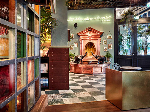 Königlicher Lobbybereich im 25hours The Royal Bavarian mit farbigen Glasflächen, Schachbrettboden, verspielter Brunneninstallation und üppiger Bepflanzung im Hintergrund.