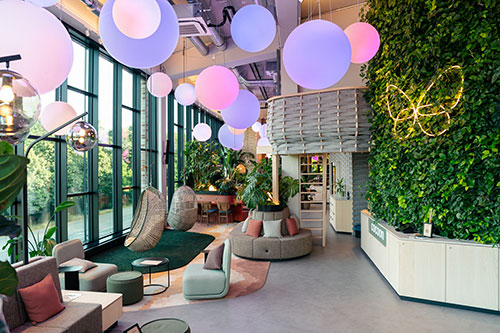 Modern gestaltete Lobby des Cocoon Theresienhöhe mit großen Panoramafenstern, hängenden Loungesesseln, Sitzinseln, vertikaler Pflanzenwand und farbig leuchtenden Kugellampen.