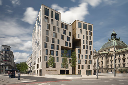 Außenansicht des neuen Hotels Koenigshof in München mit markanter moderner Fassade, großen Fensterflächen, Einschnitten im Baukörper und Blick auf das historische Justizpalast-Gebäude nebenan.