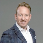 Headshot of a smiling man in a blue plaid blazer and white shirt against a gray background.