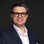 Smiling man in a navy blazer and white shirt, wearing black-framed glasses, against a dark background (professional headshot).