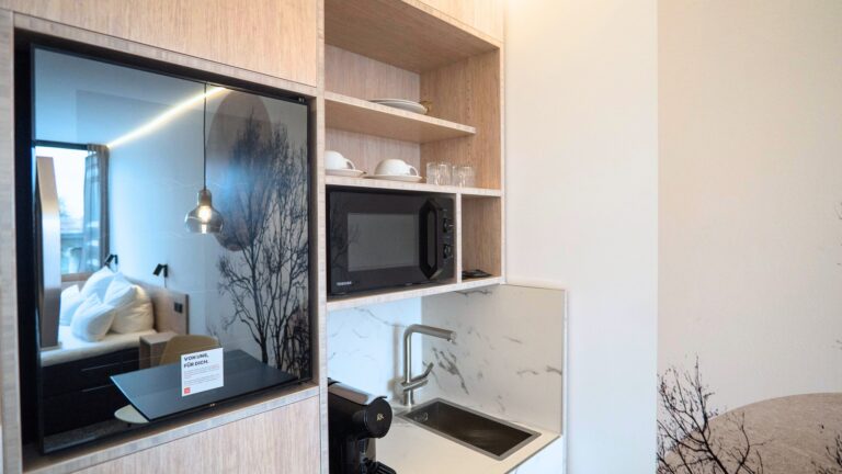 Modern kitchenette with a built-in microwave, open shelves holding dishes, and a sink, reflected bed visible in glass panel