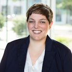 Professional headshot of a smiling woman with short hair, wearing a navy blazer and white blouse, in a bright office setting.