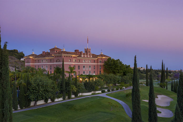 Pink courtyard hotel building with towers nestled in greenery at dusk, on a hillside overlooking a golf course path and trees.