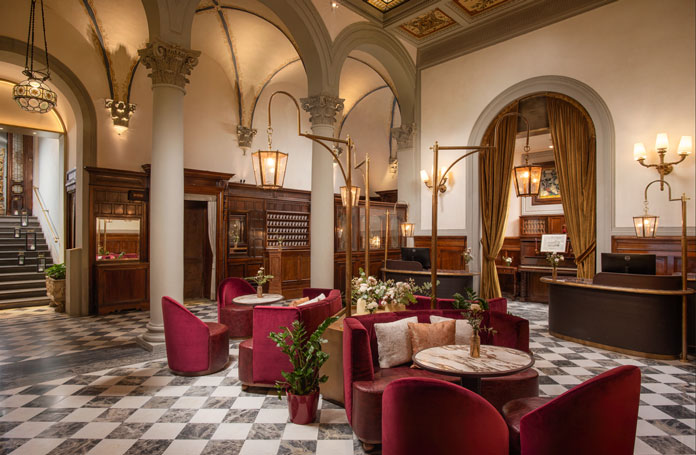 Elegant hotel lobby with a checkered marble floor, red velvet chairs around round marble tables, and arched doorways.