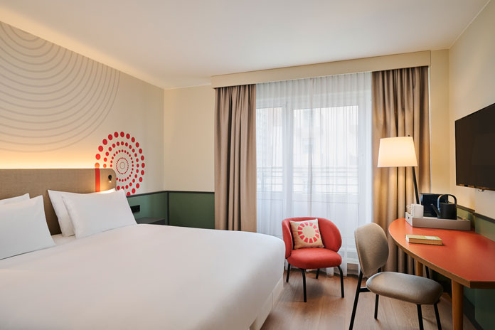 Hotel room featuring a large bed, red accent chair, desk with lamp, wall art, and a window with sheer curtains.